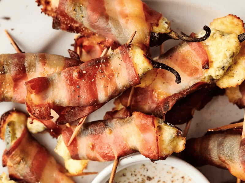 Bacon-wrapped stuffed jalape&ntilde;o peppers arranged on a plate with toothpicks, next to a small bowl of dipping sauce.