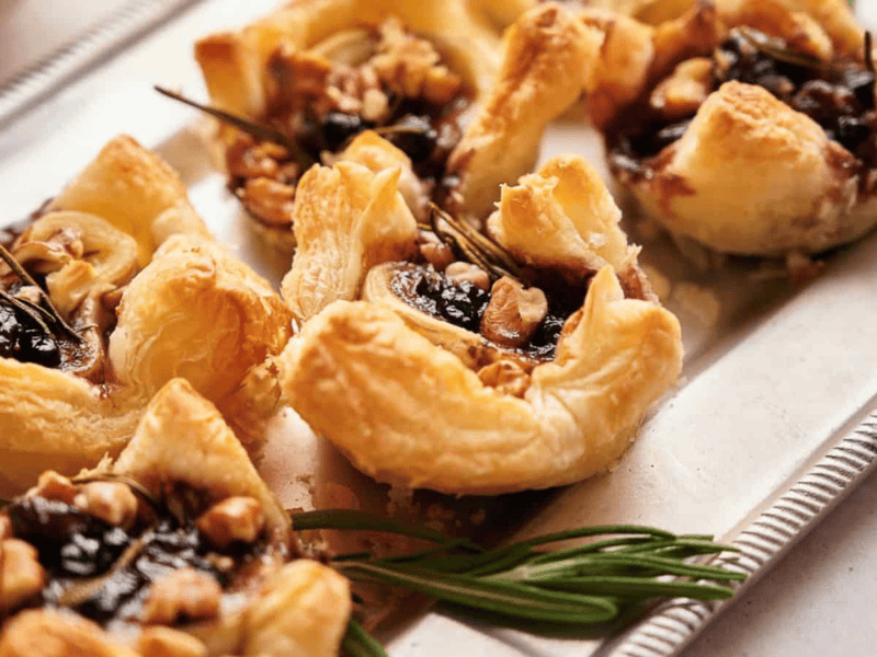 A close-up of puff pastry appetizers filled with dark jam, walnuts, and herbs, arranged on a silver tray.