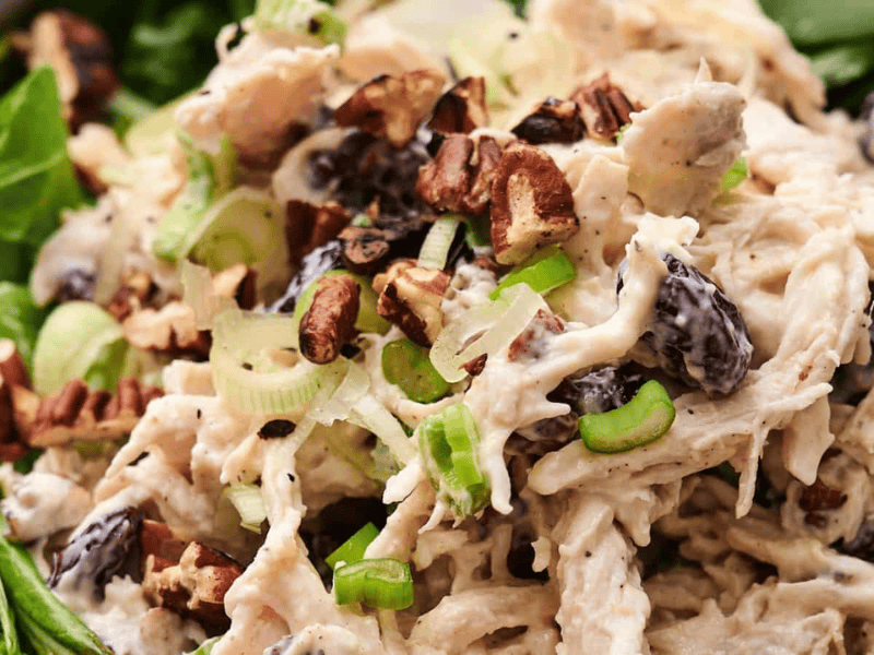 Close-up of chicken salad with shredded chicken, chopped pecans, celery slices, and dried cranberries on a bed of leafy greens.