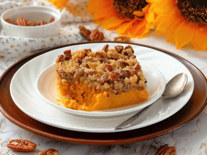 A slice of sweet potato casserole topped with pecan crumble on a white plate with a spoon, surrounded by pecans and sunflowers.