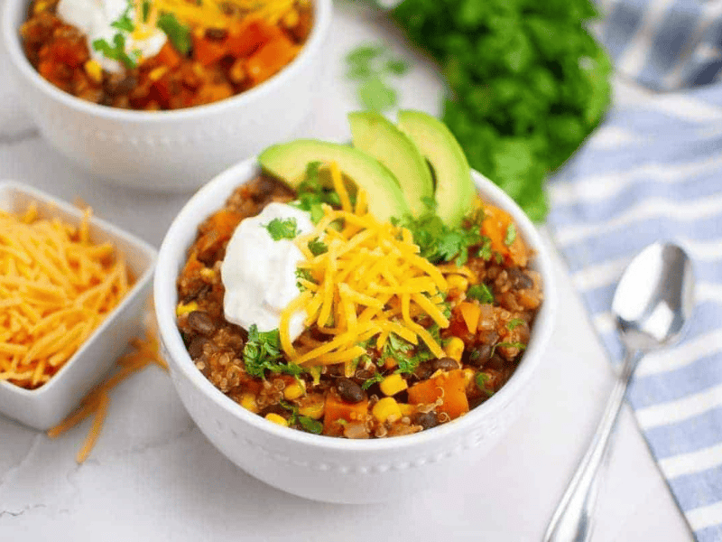 A bowl of quinoa chili topped with sliced avocado, shredded cheese, sour cream, and cilantro, with a spoon beside it and shredded cheese in a small dish nearby.