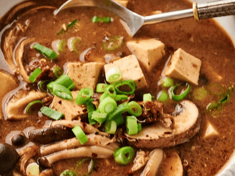 A bowl of spicy soup with tofu cubes, assorted mushrooms, sliced green onions, and a metal spoon.