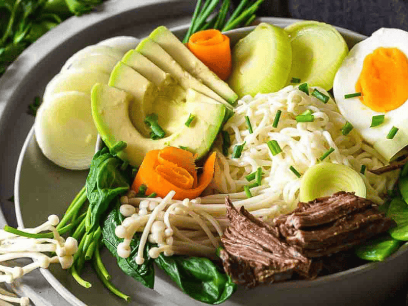 A bowl of ramen topped with sliced avocado, soft-boiled egg, shredded beef, leeks, spinach, mushrooms, and carrot slices, garnished with chopped chives.