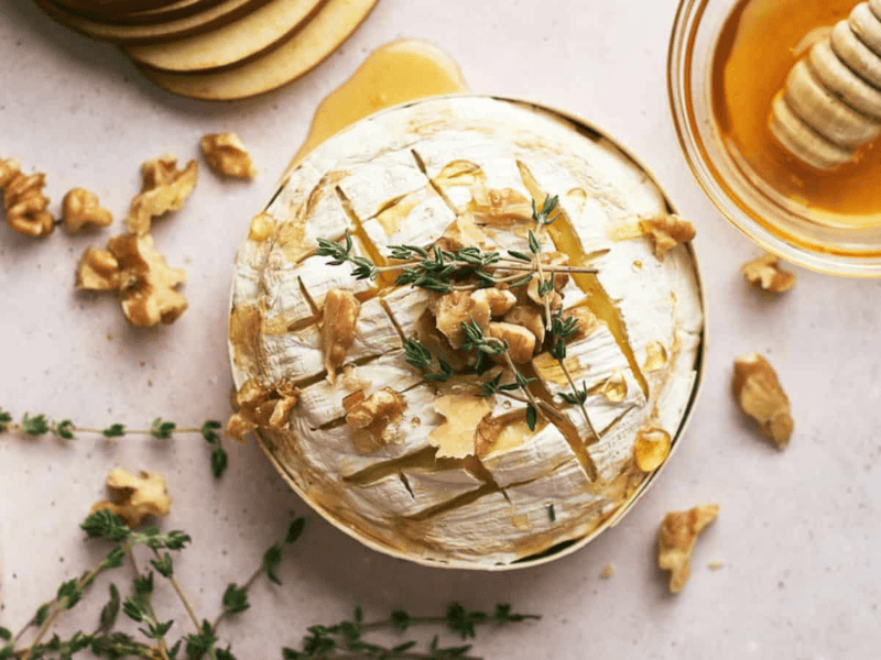 A wheel of baked brie topped with walnuts and fresh thyme, surrounded by apple slices, honey, and extra herbs on a light surface.