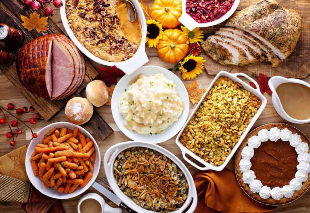 A table set with Thanksgiving foods, including turkey, ham, stuffing, mashed potatoes, gravy, green bean casserole, carrots, cranberry sauce, sweet potato casserole, pie, and rolls.