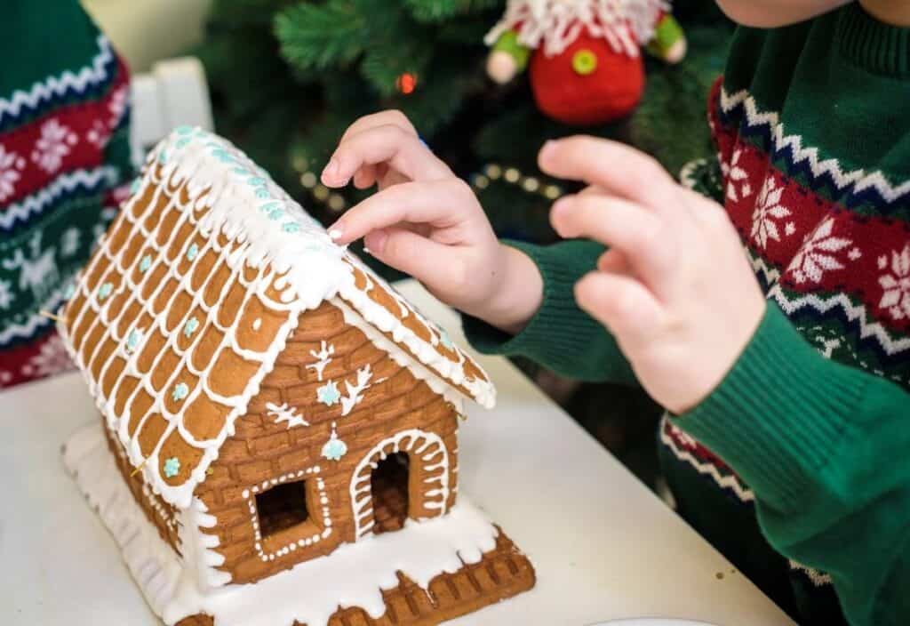 A child decorates a gingerbread house with white icing while wearing a green holiday sweater, with a Christmas tree in the background.