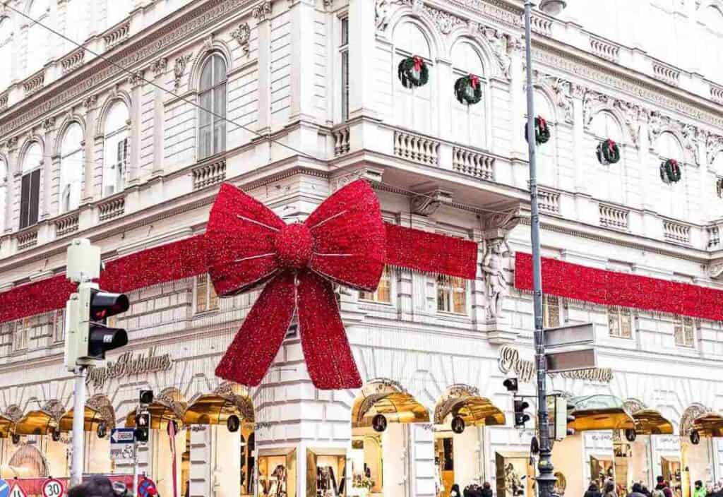 A white, ornate building decorated with a large red ribbon and bow for the holidays, with wreaths on windows and shops on the ground floor.
