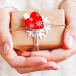 Hands holding a small gift wrapped in brown paper, tied with string, decorated with a red heart and white snowflake ornaments.