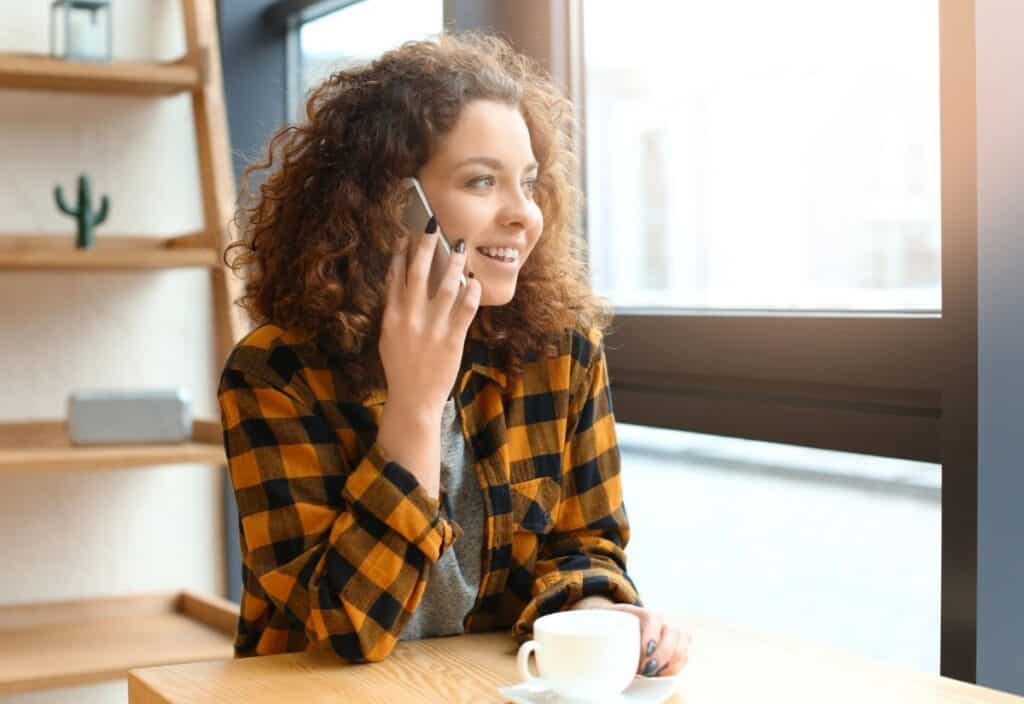 A woman with curly hair sits at a table by a window, holding a smartphone to her ear and smiling, with a white cup in front of her.