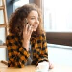 A woman with curly hair sits at a table by a window, holding a smartphone to her ear and smiling, with a white cup in front of her.