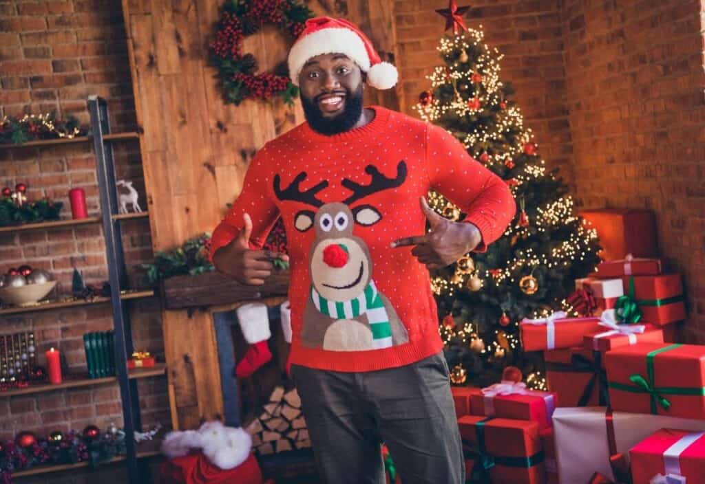 A man wearing a Santa hat and a red reindeer sweater stands indoors, smiling and pointing at his sweater in celebration of National Ugly Christmas Sweater Day, with a decorated Christmas tree and wrapped gifts in the background.