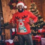 A man wearing a Santa hat and a red reindeer sweater stands indoors, smiling and pointing at his sweater in celebration of National Ugly Christmas Sweater Day, with a decorated Christmas tree and wrapped gifts in the background.