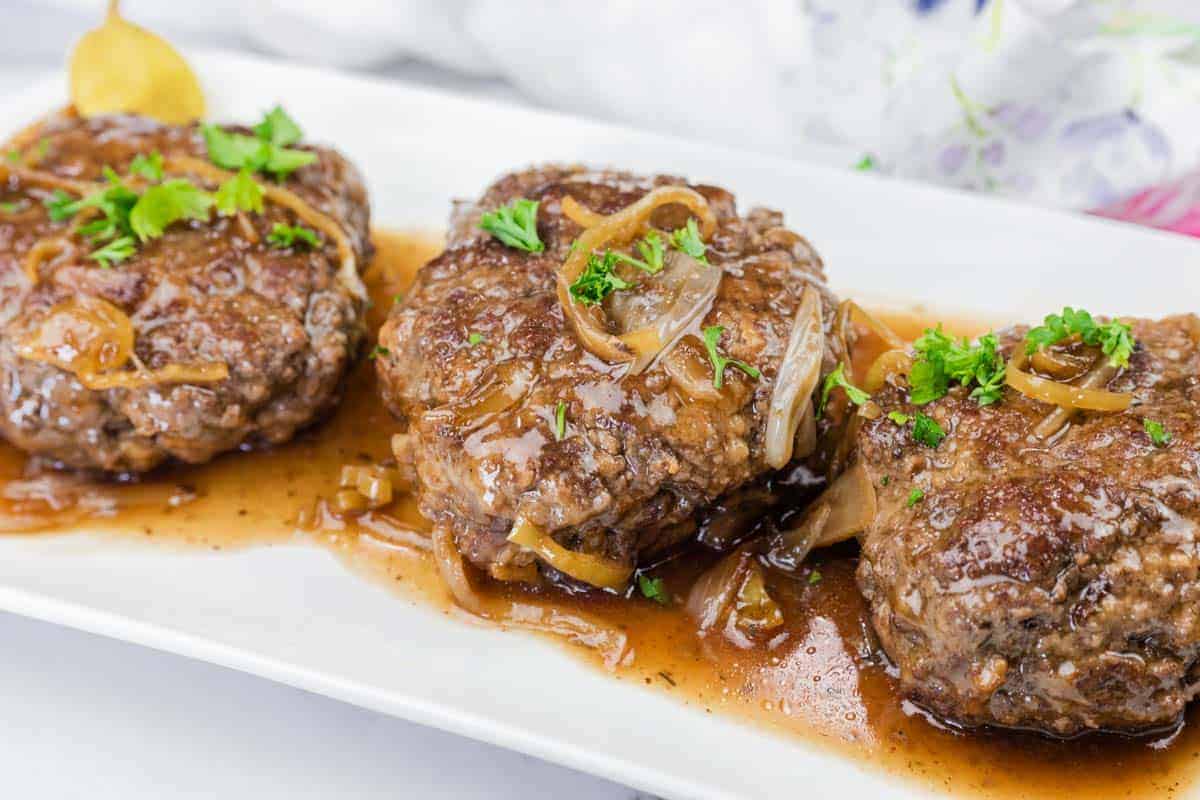 Three Mississippi Salisbury Steak covered in brown onion gravy, garnished with chopped parsley, served on a white rectangular plate.