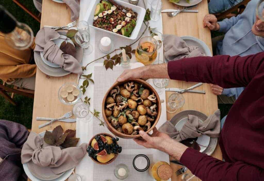 People sit around a wooden table set for a meal, as one person serves a bowl of roasted potatoes and mushrooms. Other dishes and drinks are arranged on the table.