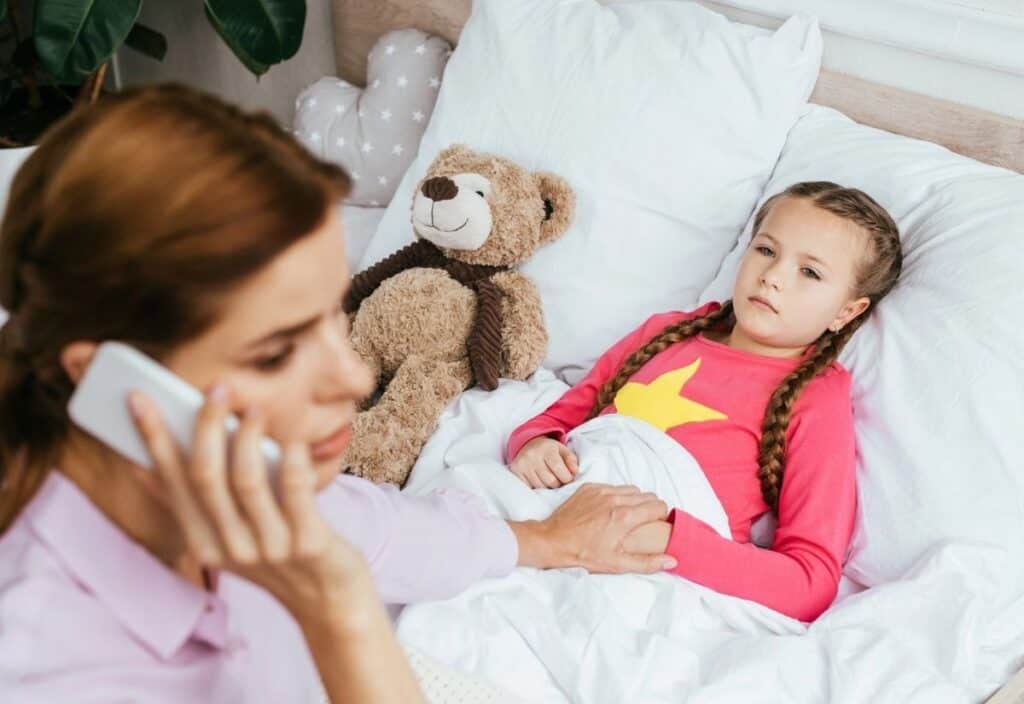 A woman talks on the phone while holding the hand of a young girl lying in bed, who looks unwell. A teddy bear is next to the girl.