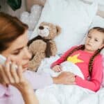 A woman talks on the phone while holding the hand of a young girl lying in bed, who looks unwell. A teddy bear is next to the girl.