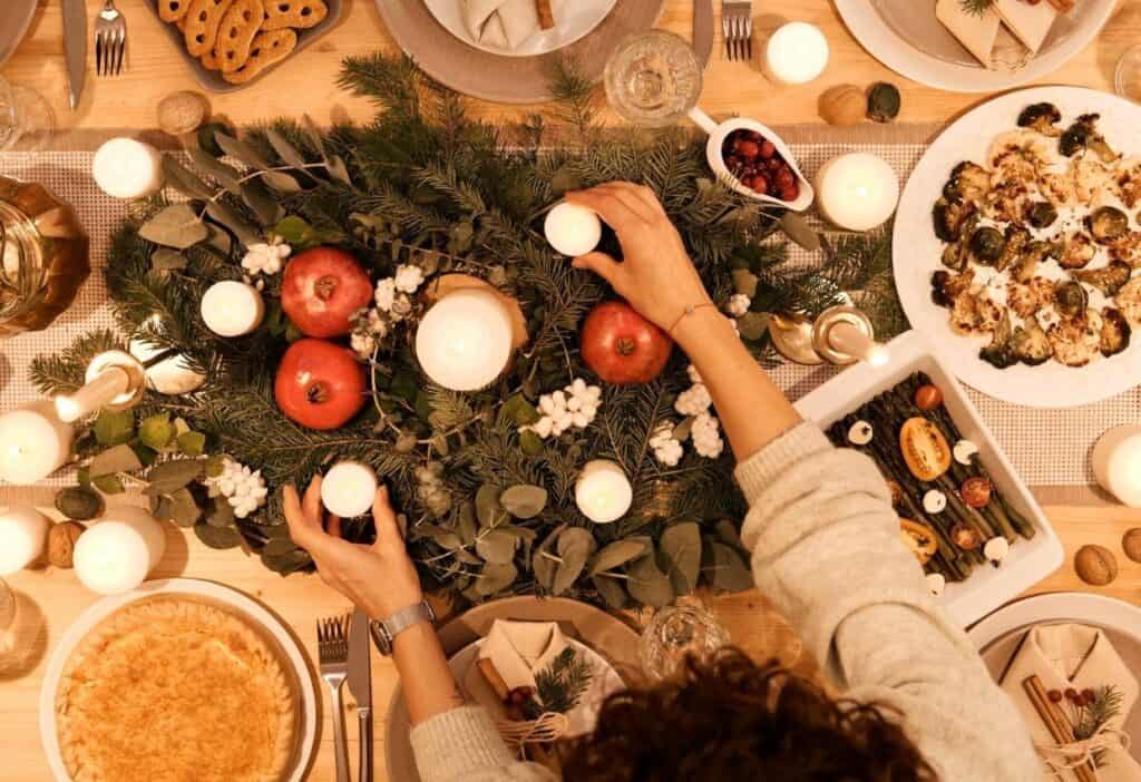 Person arranging small candles on a holiday dinner table decorated with greenery, pomegranates, and festive dishes for a cozy meatless Christmas feast. Plates of food and drinks surround the elegant centerpiece.