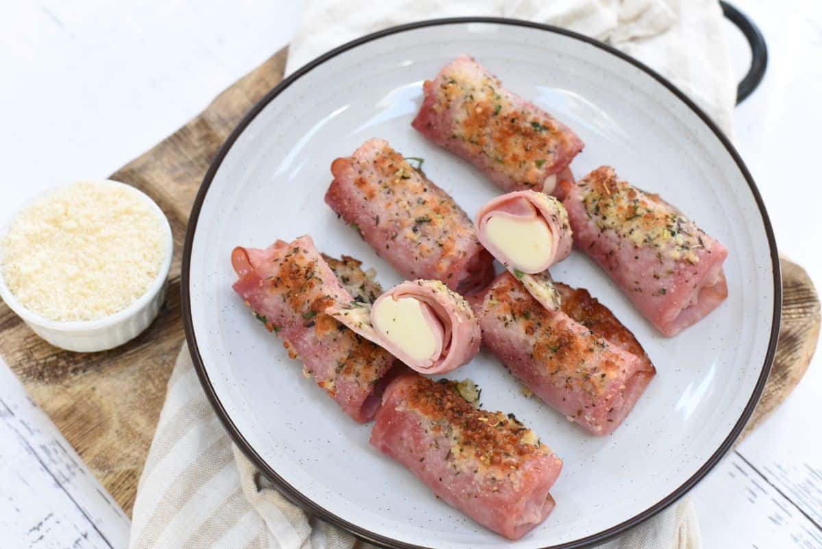 A plate with seven ham rolls filled with cheese, some cut to show the filling, next to a small bowl of grated cheese on a wooden board.