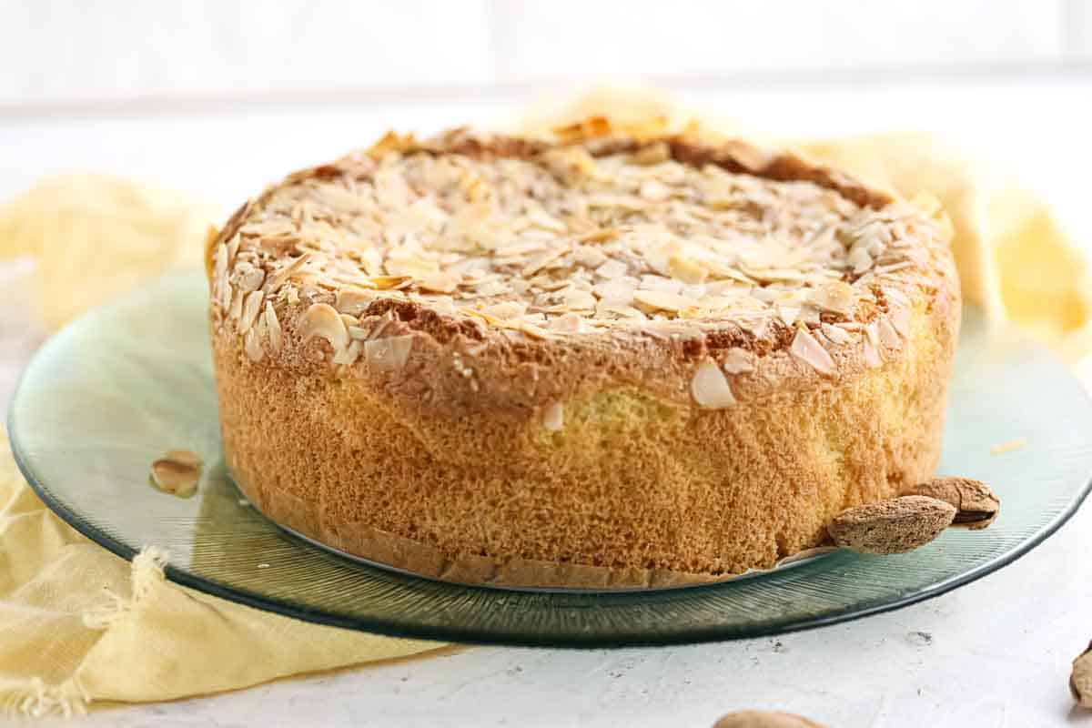 A round almond cake topped with sliced almonds sits on a glass plate, with a yellow cloth and whole almonds nearby.