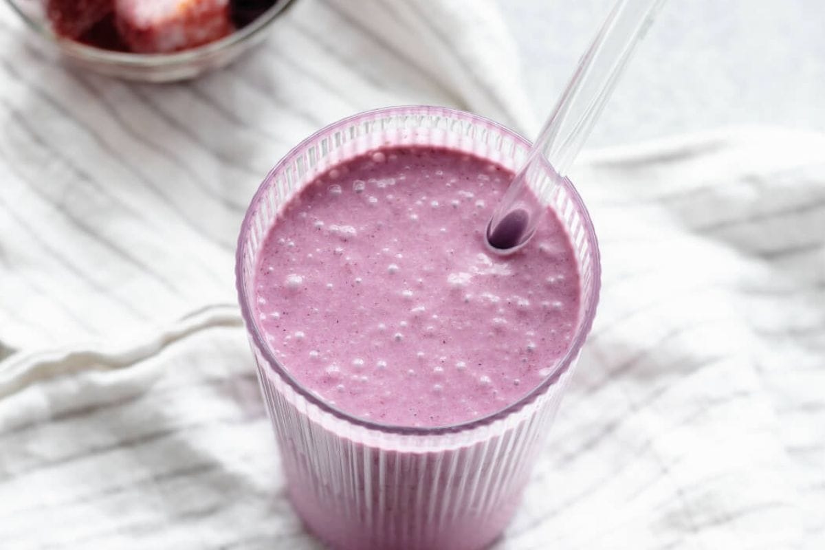 A glass of thick, purple smoothie with a clear straw, placed on a white cloth with a bowl of strawberries in the background.