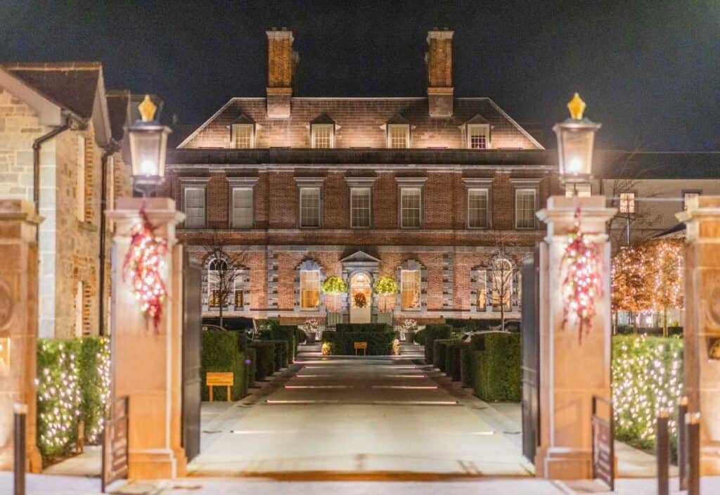 A large, stately brick mansion is illuminated at night, with holiday lights and wreaths decorating the entrance and stone pillars in the foreground.