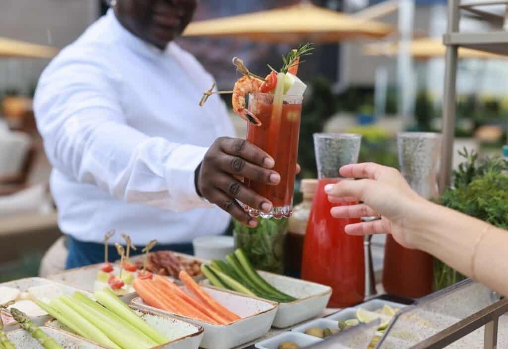 A person hands a Bloody Mary cocktail, garnished with shrimp and vegetables, to another person at an outdoor food station with fresh vegetables and drink ingredients.