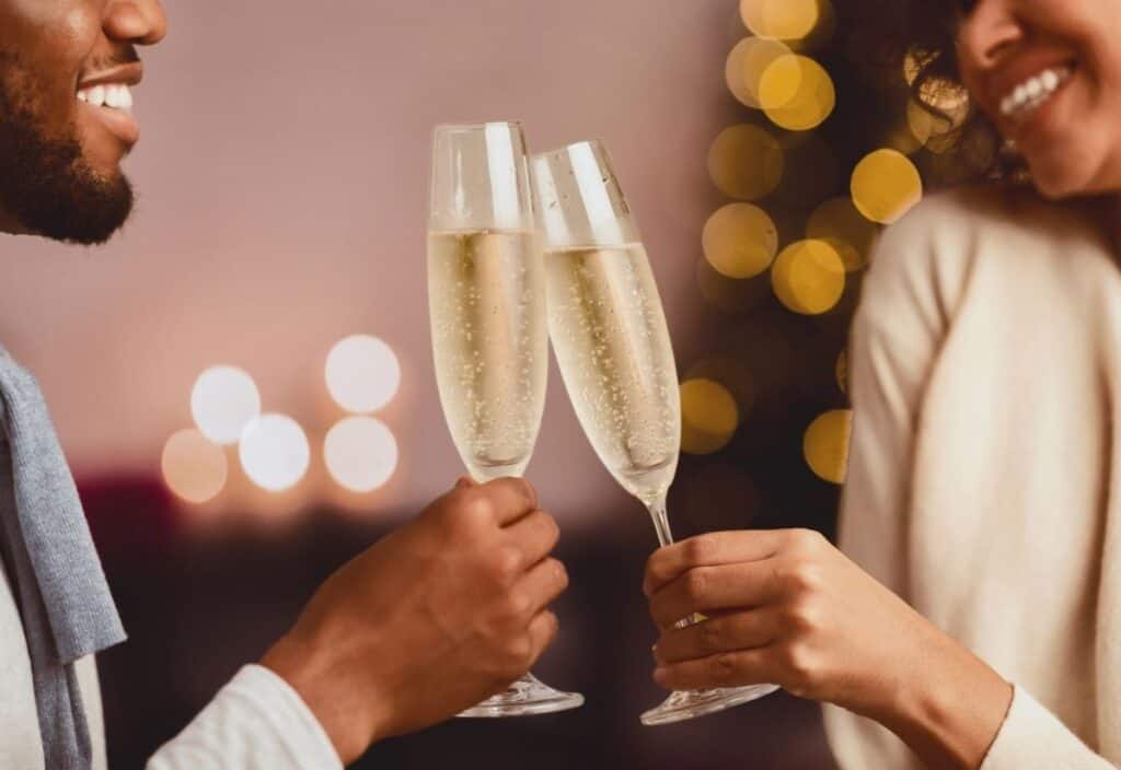 Two people clinking champagne glasses, smiling, with blurred lights and a festive background, celebrate National Champagne Day in style.