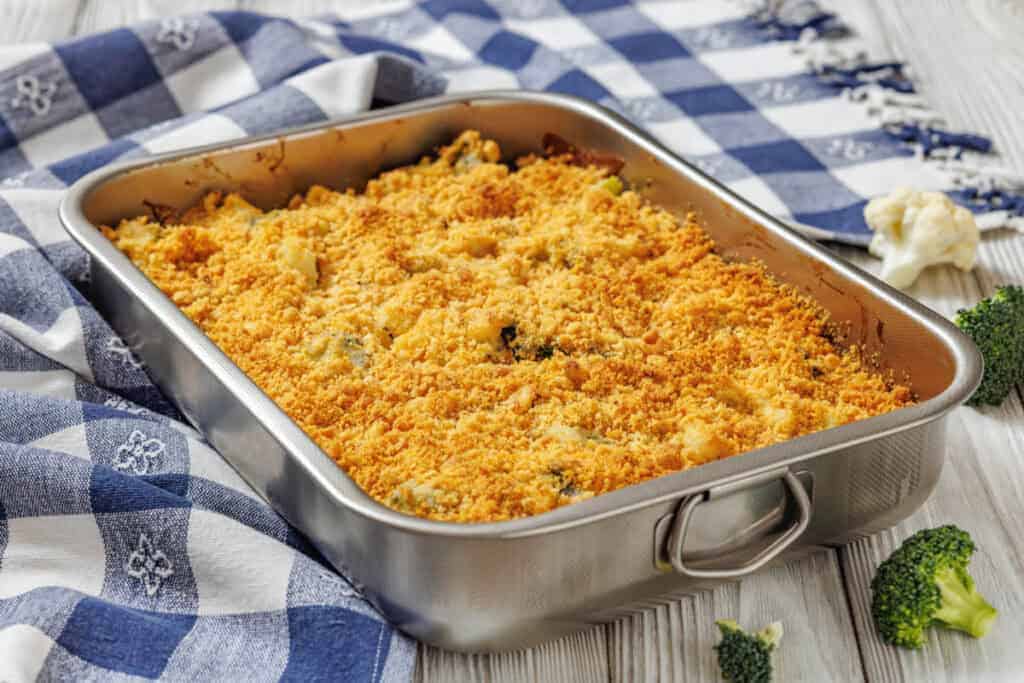 Cheesy Broccoli & Cauliflower Casserole with Crispy Bacon in a metal dish on a blue and white checkered cloth, with broccoli and cauliflower nearby.