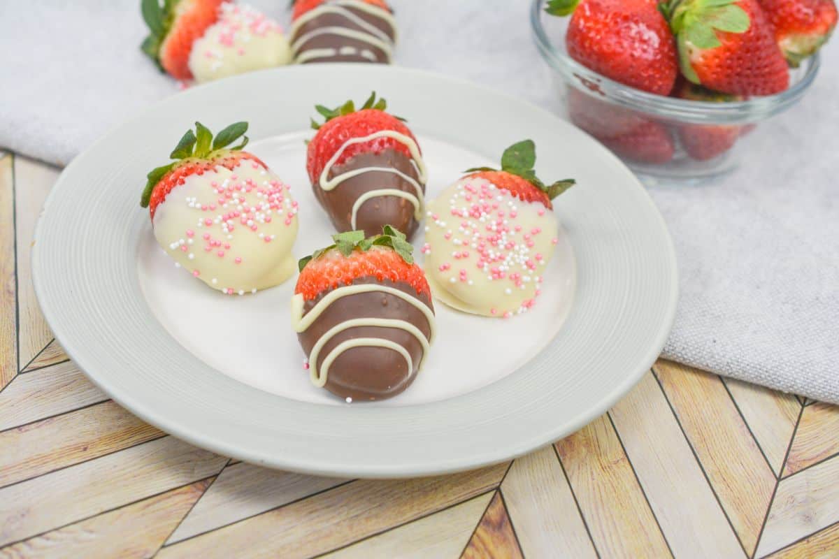 A plate of strawberries dipped in white and milk chocolate, decorated with sprinkles, sits on a wooden table next to a bowl of fresh strawberries.