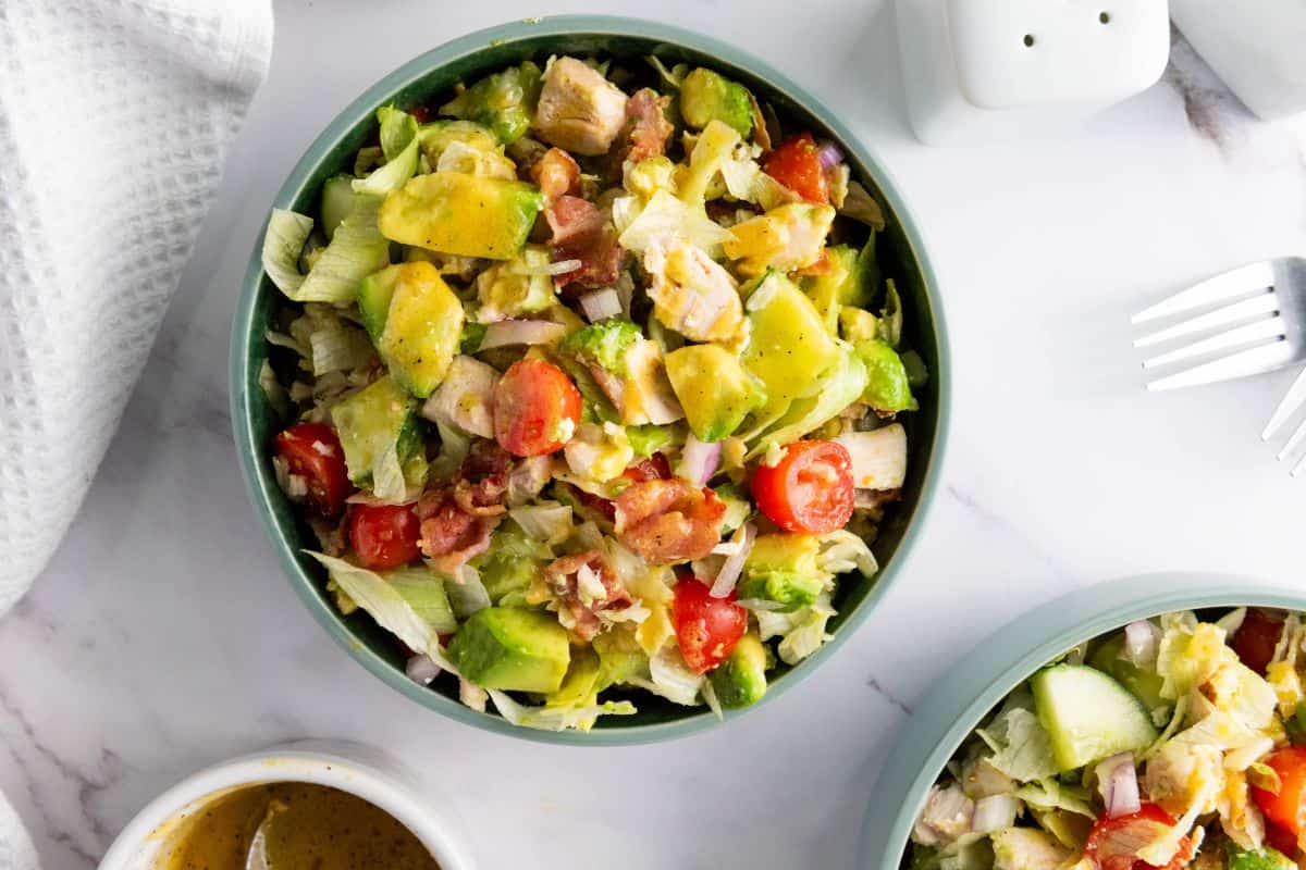 A bowl of chopped salad with lettuce, avocado, cherry tomatoes, red onion, chicken, and bacon on a white surface.