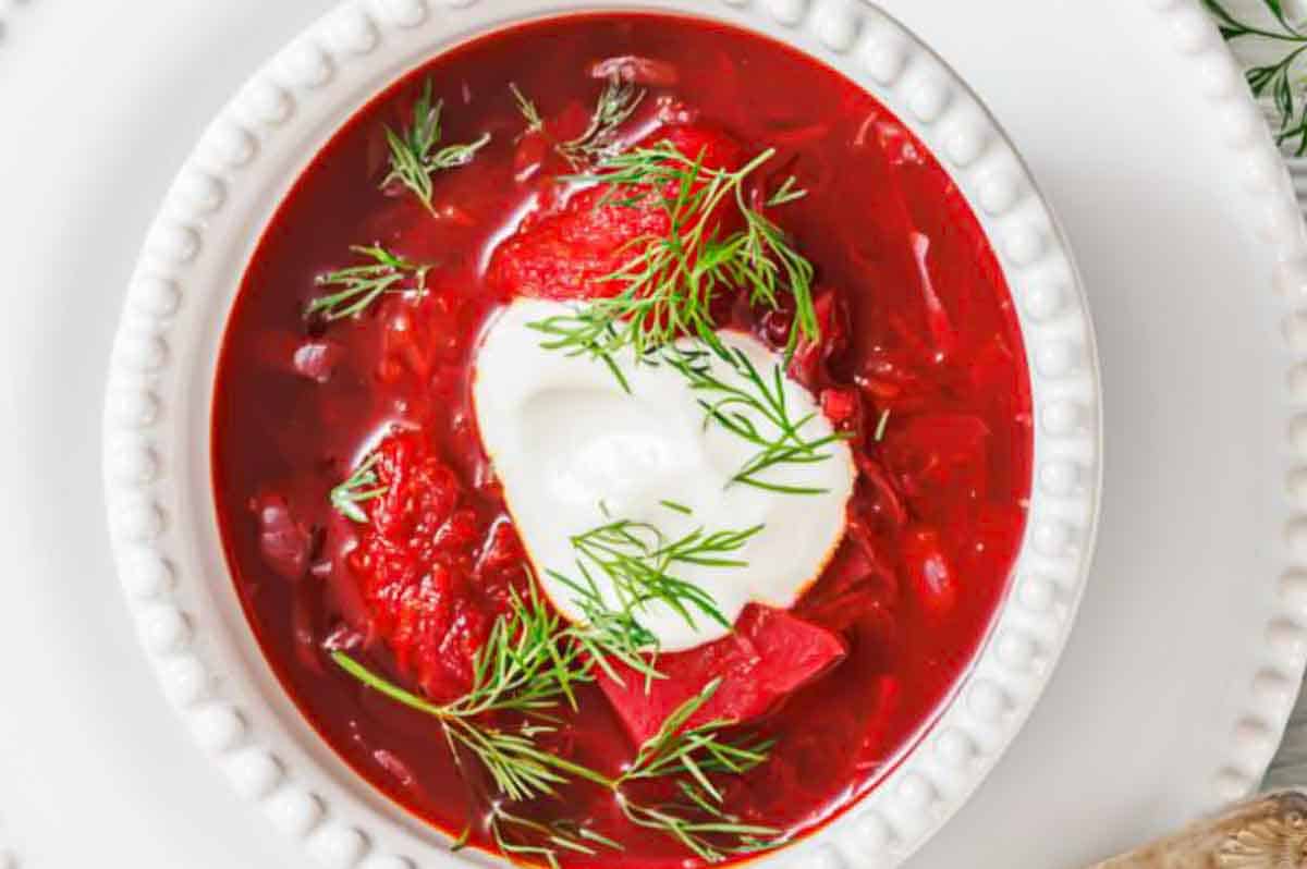 A bowl of red borscht soup topped with a dollop of sour cream and fresh dill, served in a white textured bowl.