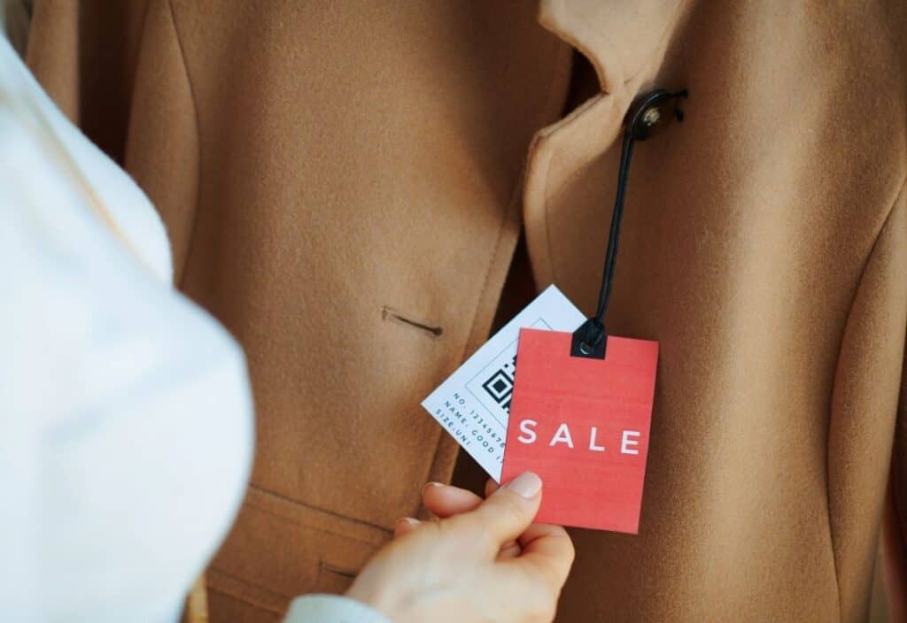 A person holds a red sale tag attached to a tan coat on display, with another tag showing a QR code in the background.