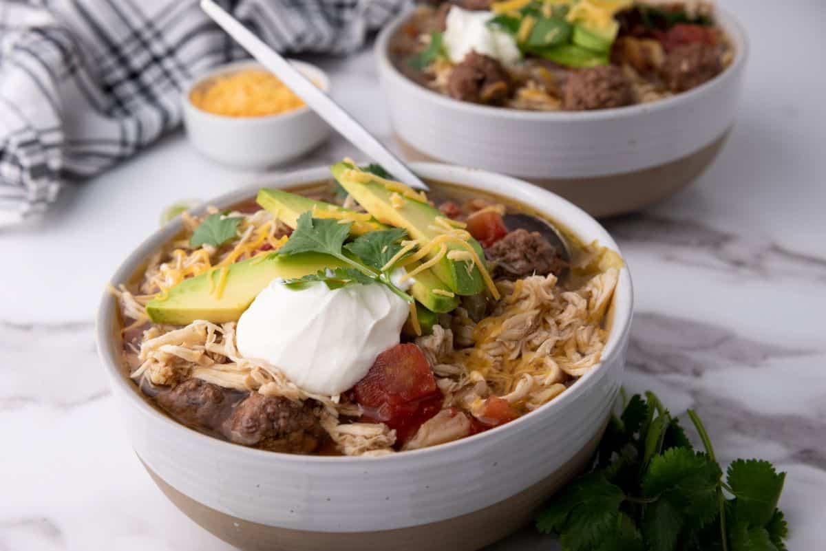 A bowl of shredded chicken and beef chili topped with sour cream, sliced avocado, cheese, and cilantro, with another similar bowl in the background.