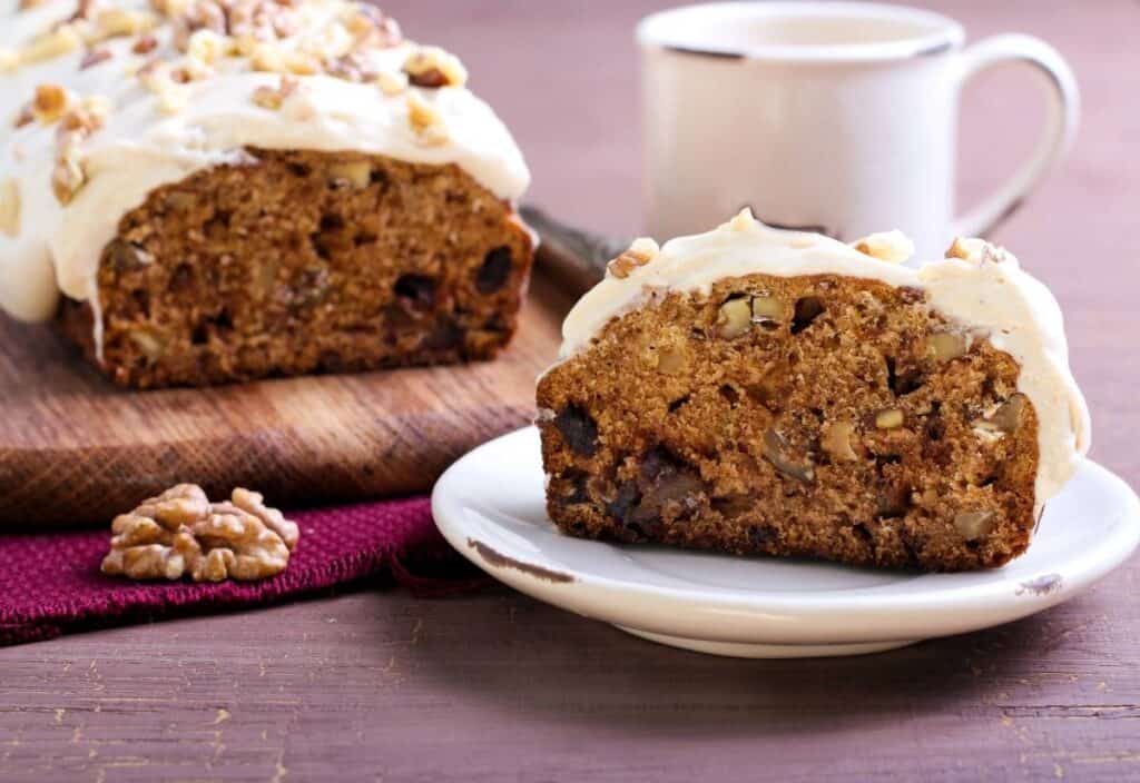 A slice of frosted nut bread on a plate, with the remaining loaf and a mug in the background on a wooden table&mdash;perfect for celebrating National Date Nut Bread Day.