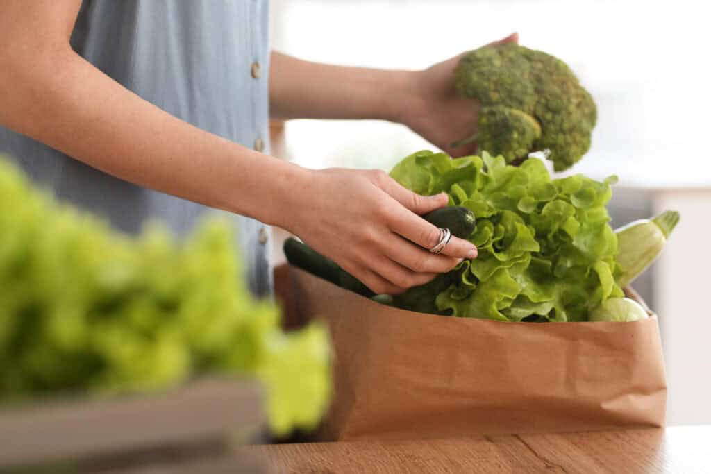 Person holding broccoli and unpacking fresh vegetables, including lettuce and cucumber, from a brown paper bag on a wooden table.