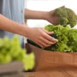 Person holding broccoli and unpacking fresh vegetables, including lettuce and cucumber, from a brown paper bag on a wooden table.