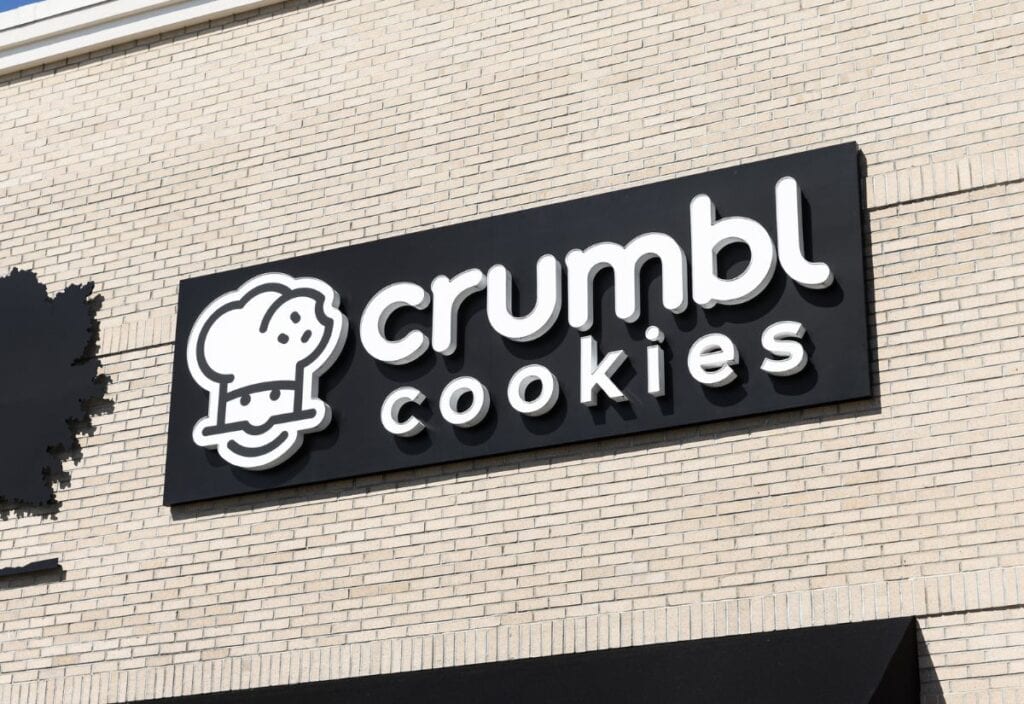 A Crumbl Cookies store sign with a chef's hat logo on a beige brick wall, reflecting the rise and fall of Crumbl Cookies in the competitive dessert market.