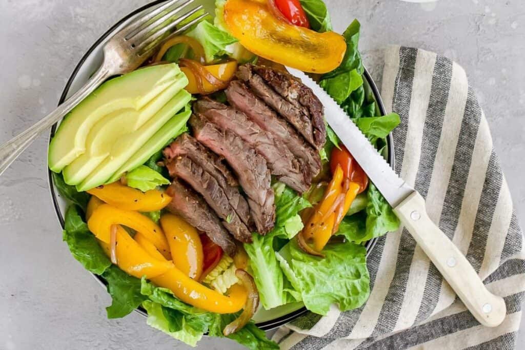 A bowl of salad with sliced steak, avocado, yellow bell peppers, and leafy greens, served with a fork and knife on a striped napkin.