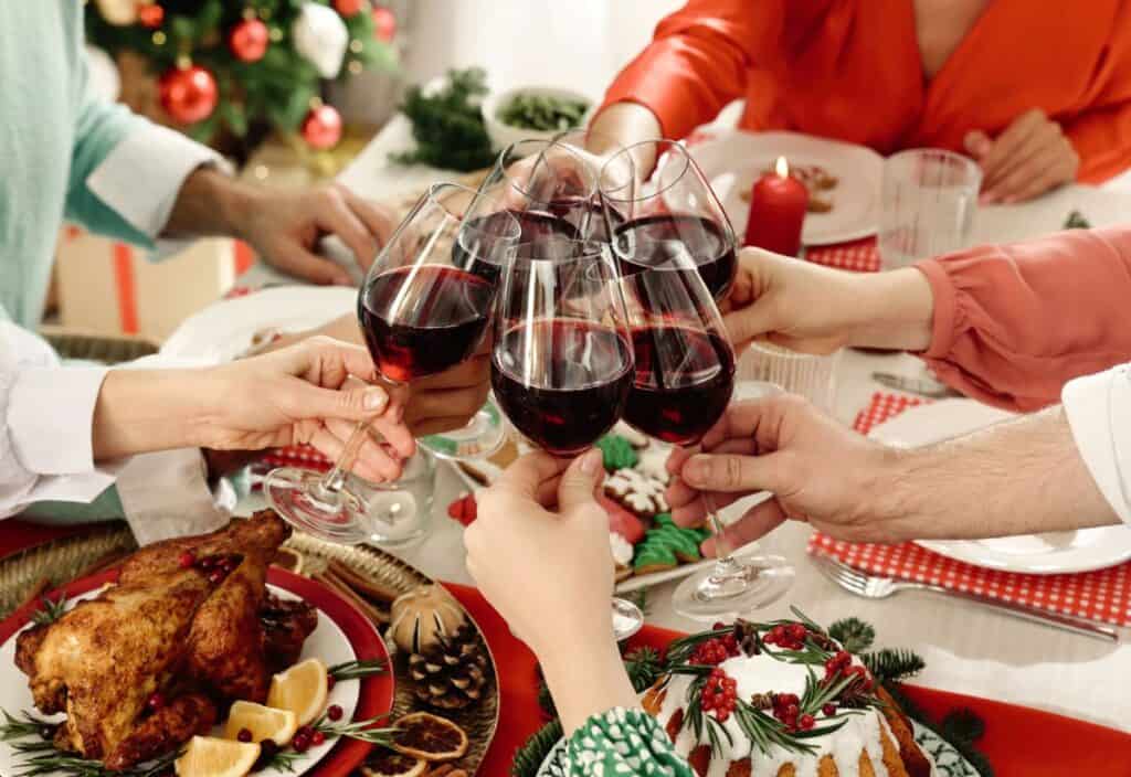 Several people clink glasses of red wine over a festive holiday dinner table decorated with food, greenery, and candles.