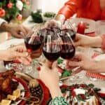 Several people clink glasses of red wine over a festive holiday dinner table decorated with food, greenery, and candles.