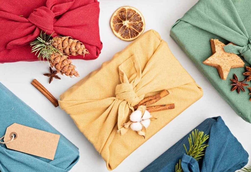 Five fabric-wrapped gifts are arranged on a table, each decorated with natural elements like pinecones, cinnamon sticks, dried orange, and evergreen sprigs&mdash;showcasing a beautiful way to reduce holiday waste.