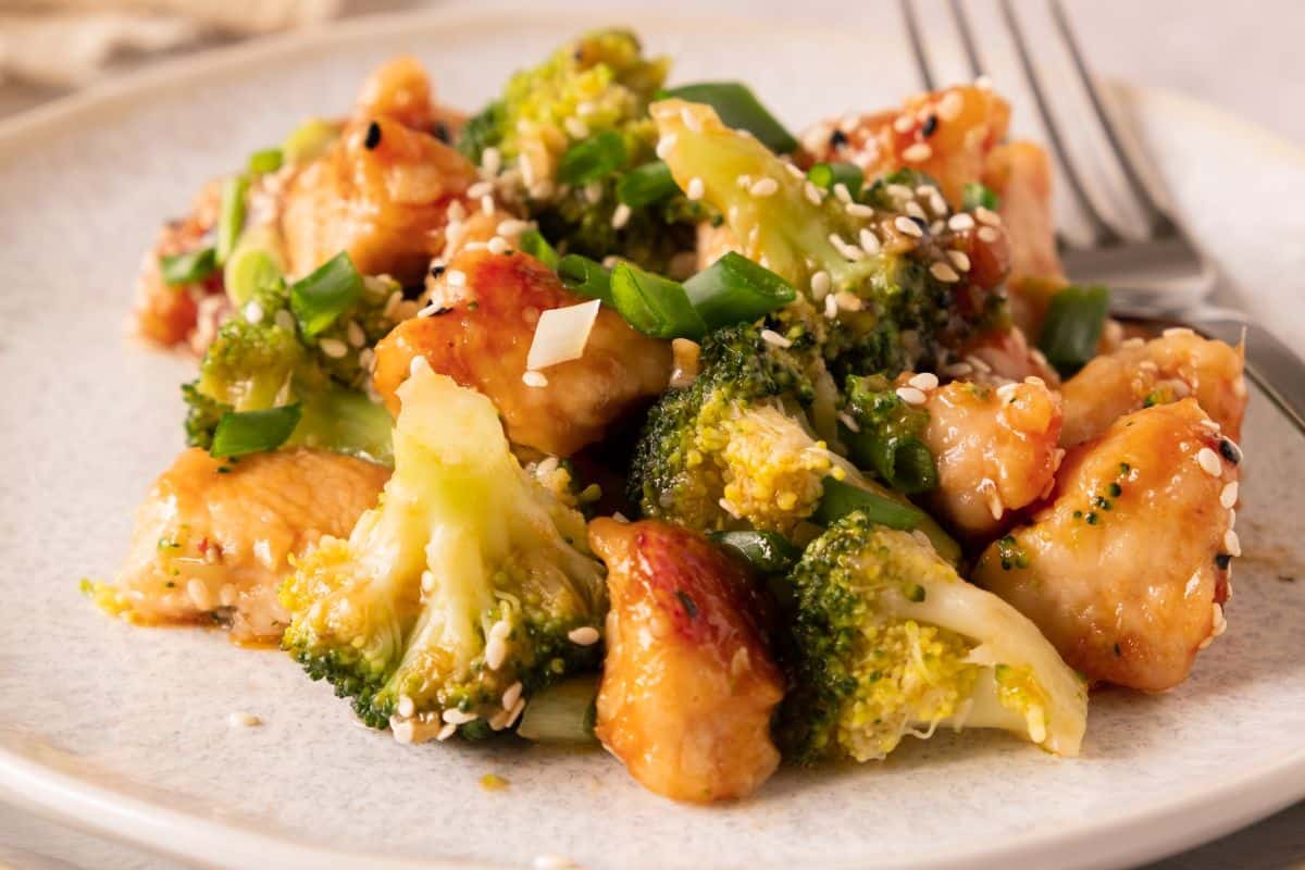 A plate of stir-fried chicken and broccoli garnished with sesame seeds and chopped green onions, with a fork beside the food.