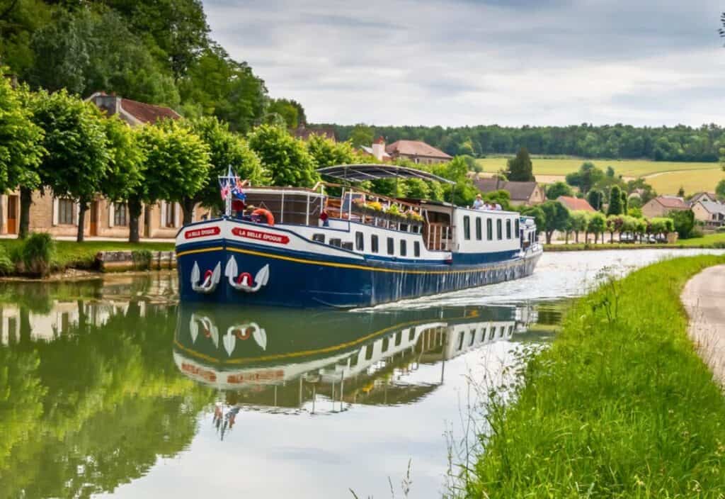 A blue and white canal barge glides along a calm, tree-lined waterway, offering a luxury barge cruise past rural houses and green fields in the background.