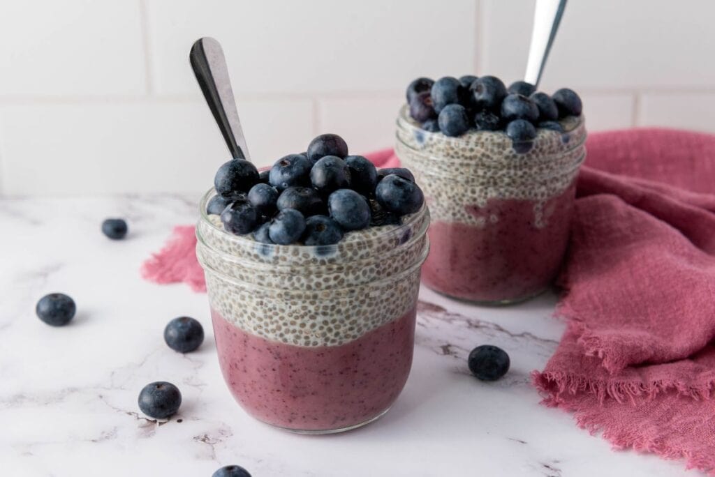 Two glass jars filled with layered chia pudding and topped with fresh blueberries sit on a marble surface, with spoons and a pink cloth nearby.