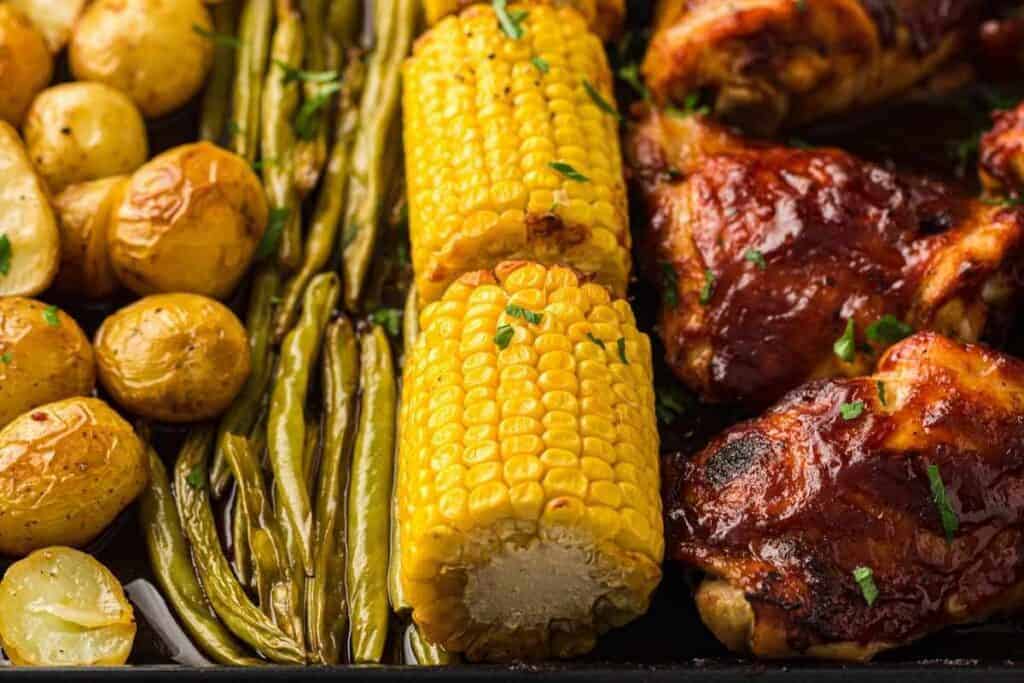 Roasted baby potatoes, green beans, corn on the cob, and barbecue chicken arranged on a sheet pan.