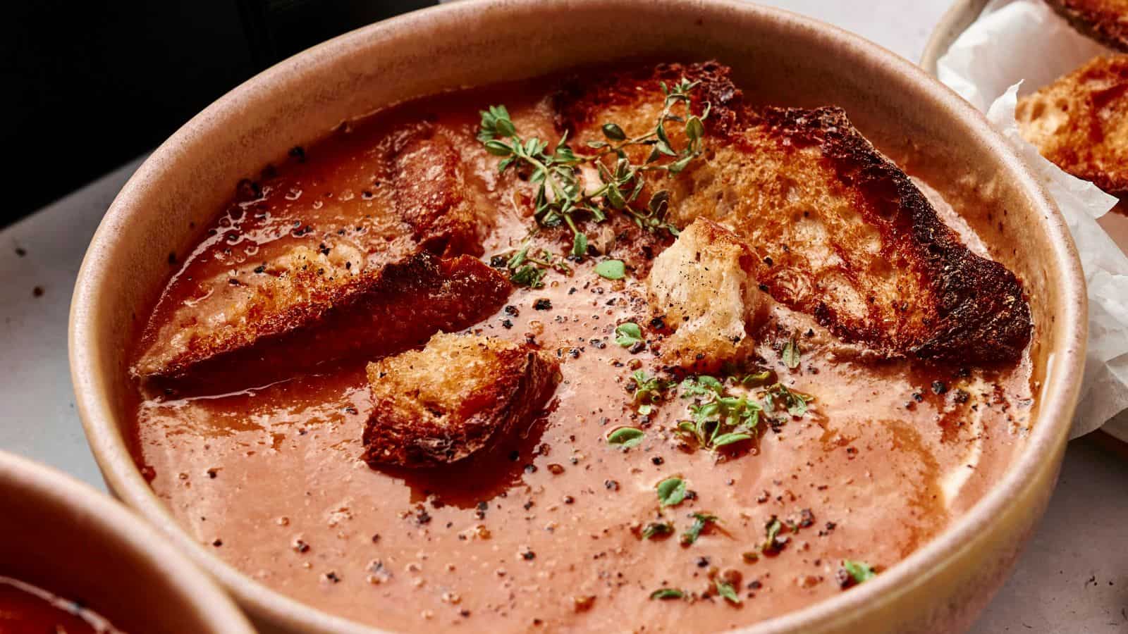 A bowl of tomato soup topped with toasted bread pieces, fresh thyme sprigs, and cracked black pepper.
