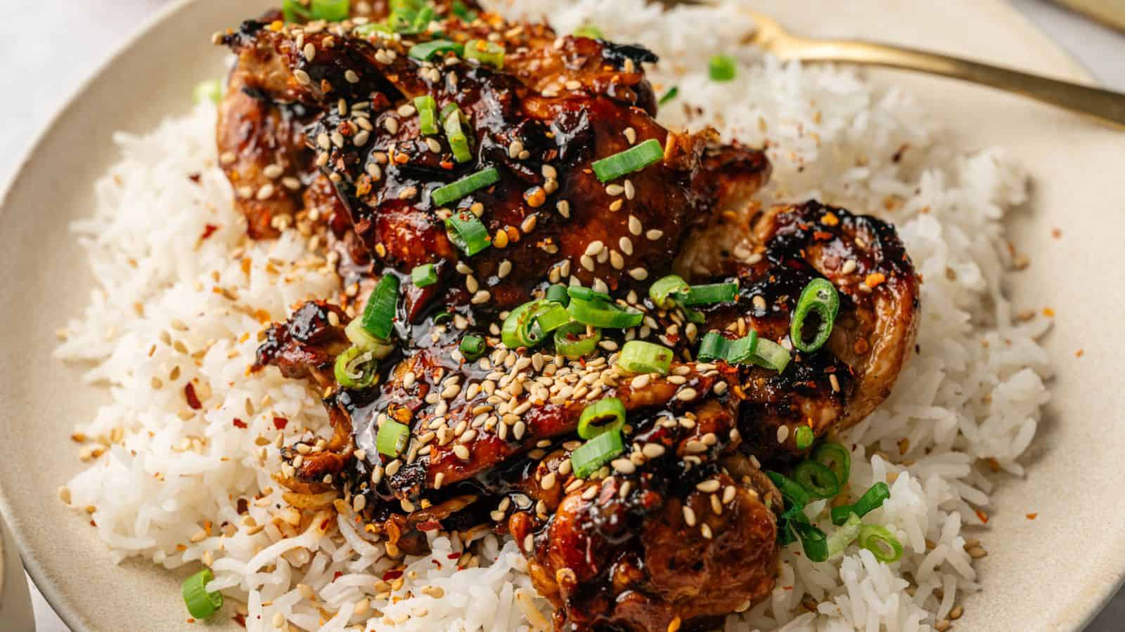 A plate of white rice topped with glazed chicken pieces, garnished with sesame seeds and chopped green onions.