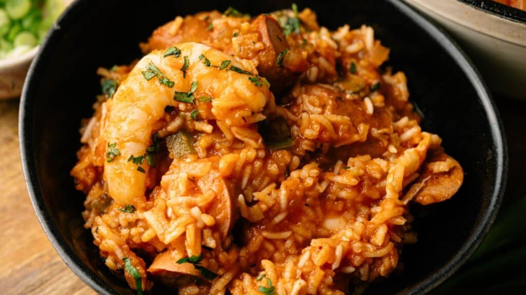 A black bowl filled with jambalaya, featuring shrimp, sausage, rice, and vegetables in a reddish sauce, garnished with chopped herbs.