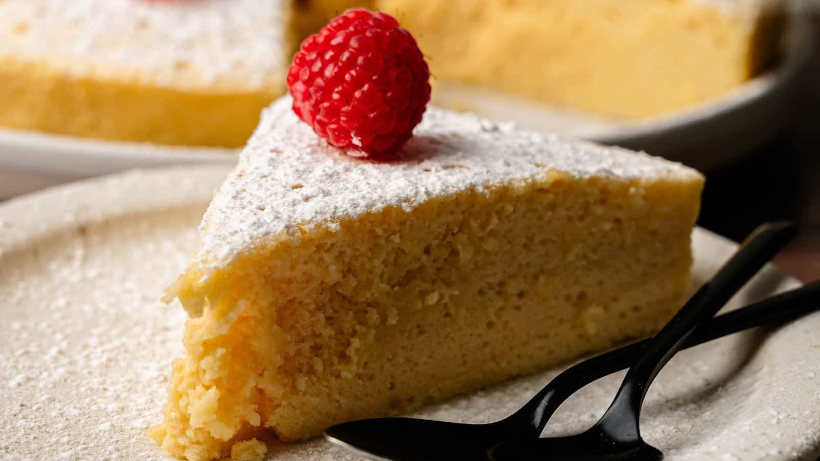 A slice of sponge cake topped with powdered sugar and a raspberry, served on a plate with two black plastic forks.