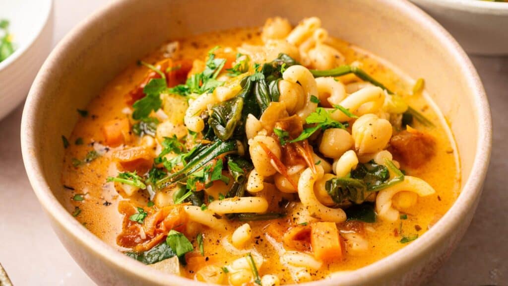 A bowl of pasta soup with chickpeas, spinach, tomatoes, and herbs in a creamy broth.